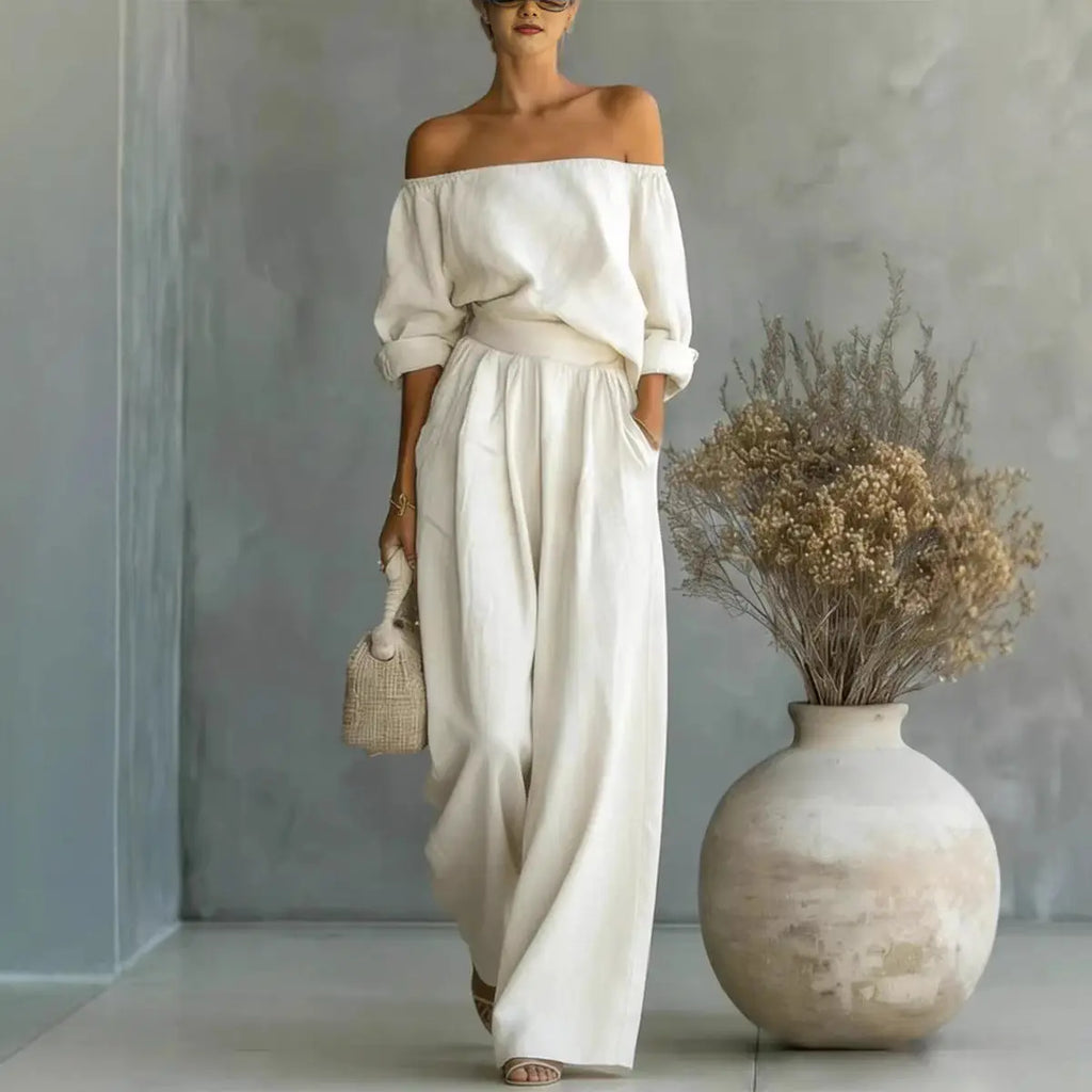 A woman models an off-the-shoulder cream linen jumpsuit with wide-leg pants, standing in a minimalist room with a textured gray wall and a large ceramic vase with dried flowers.