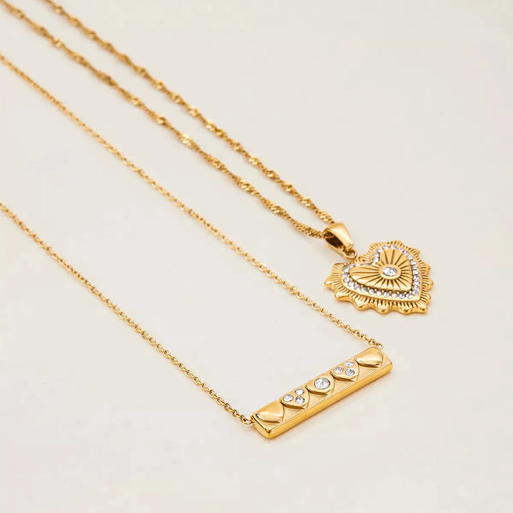 Two gold necklaces with heart and bar pendants on a light beige background