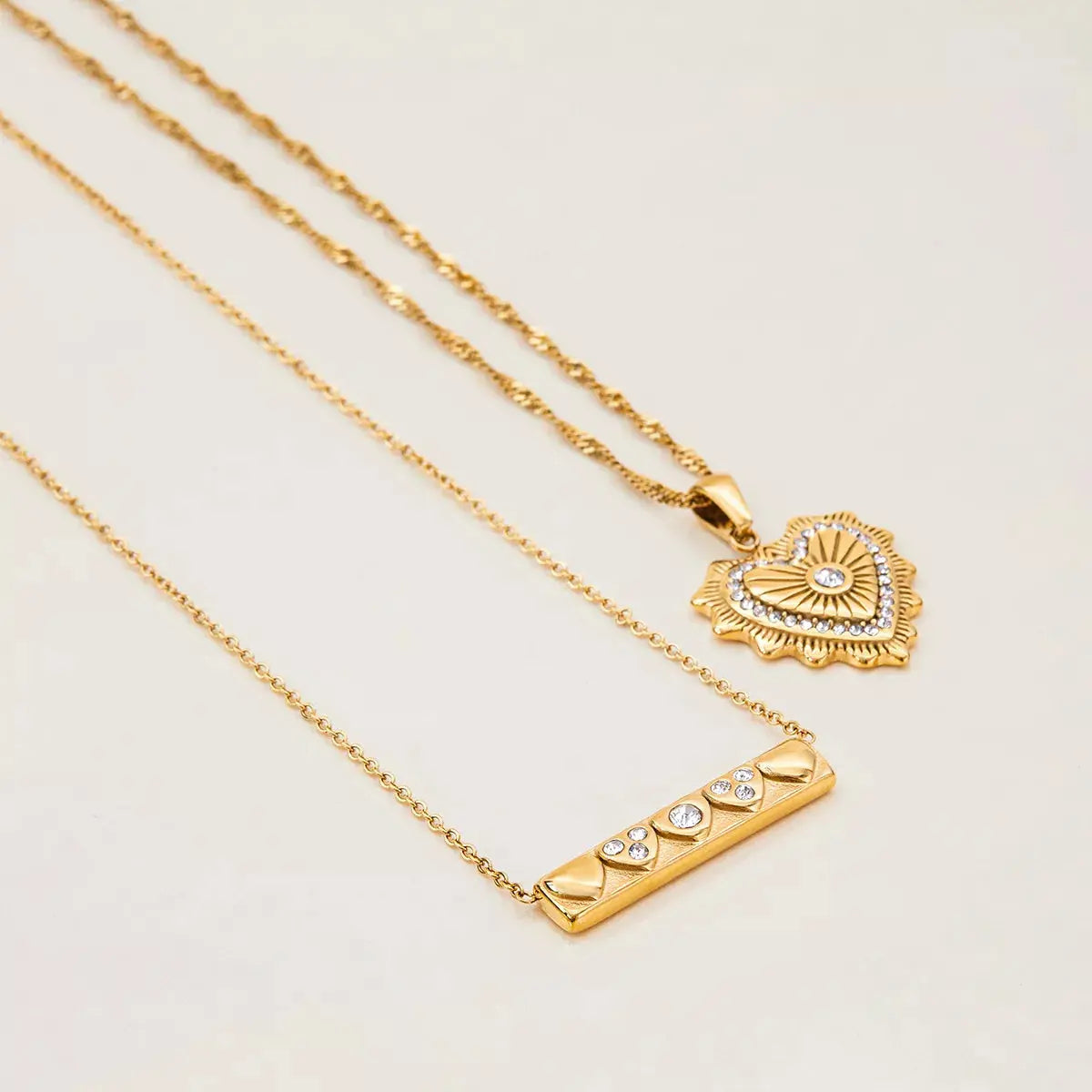 Two gold necklaces with heart and bar pendants on a light beige background
