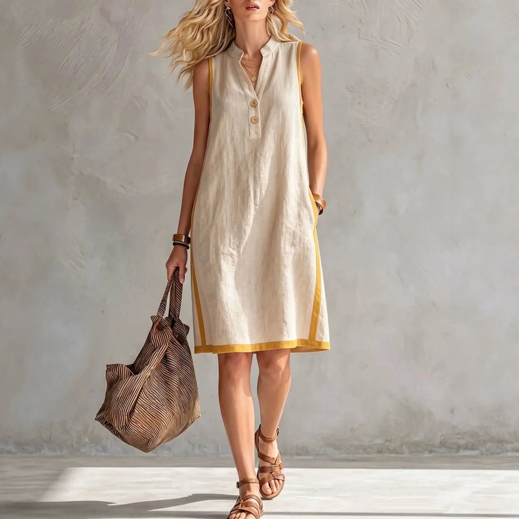 Woman wearing a beige sleeveless dress with yellow trim, holding a brown bag against a plain background.