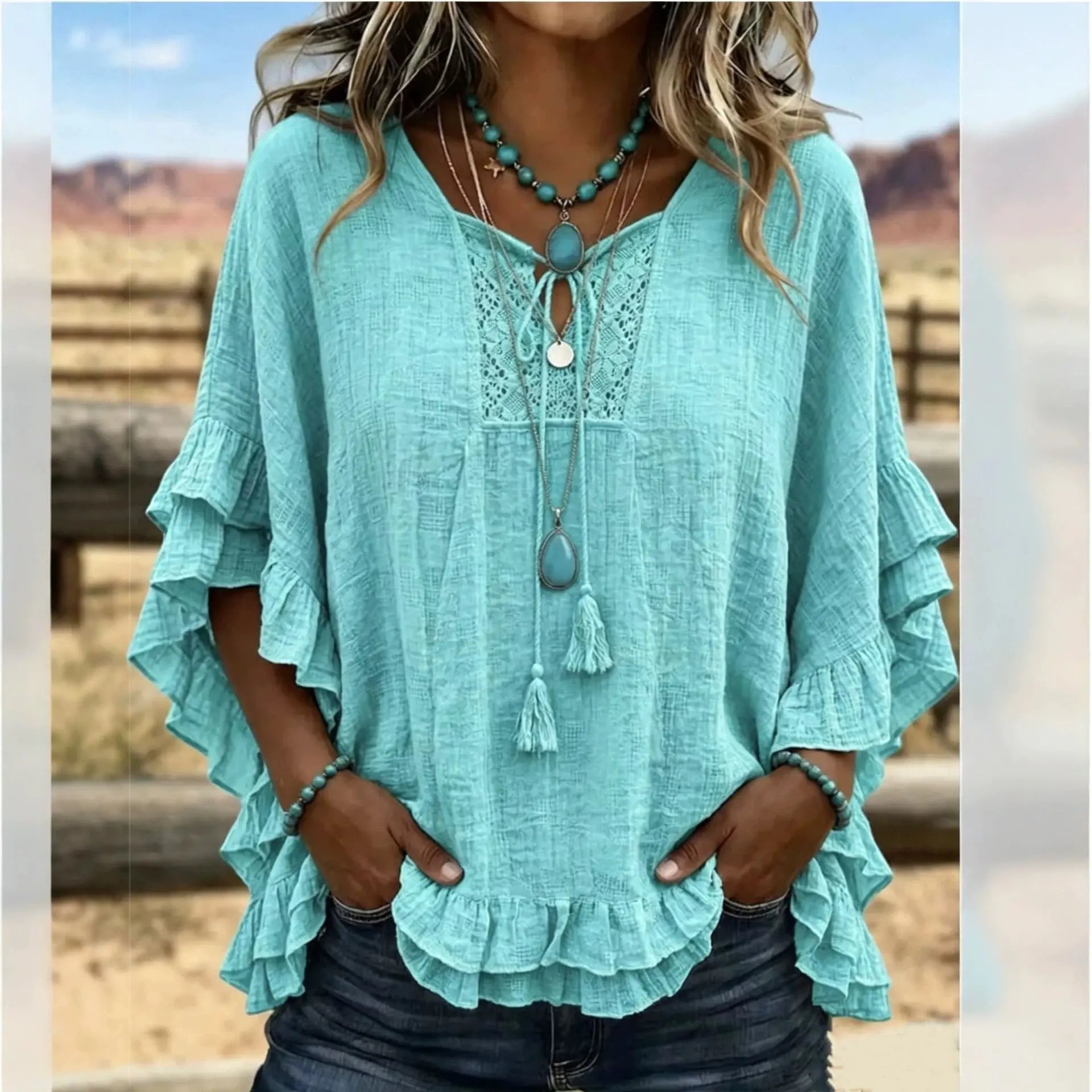 A woman wearing a light aqua blue bohemian-style blouse with layered ruffle sleeves, a lace V-neckline, and a tassel tie, paired with dark jeans and turquoise jewelry, set against a blurred outdoor desert background.