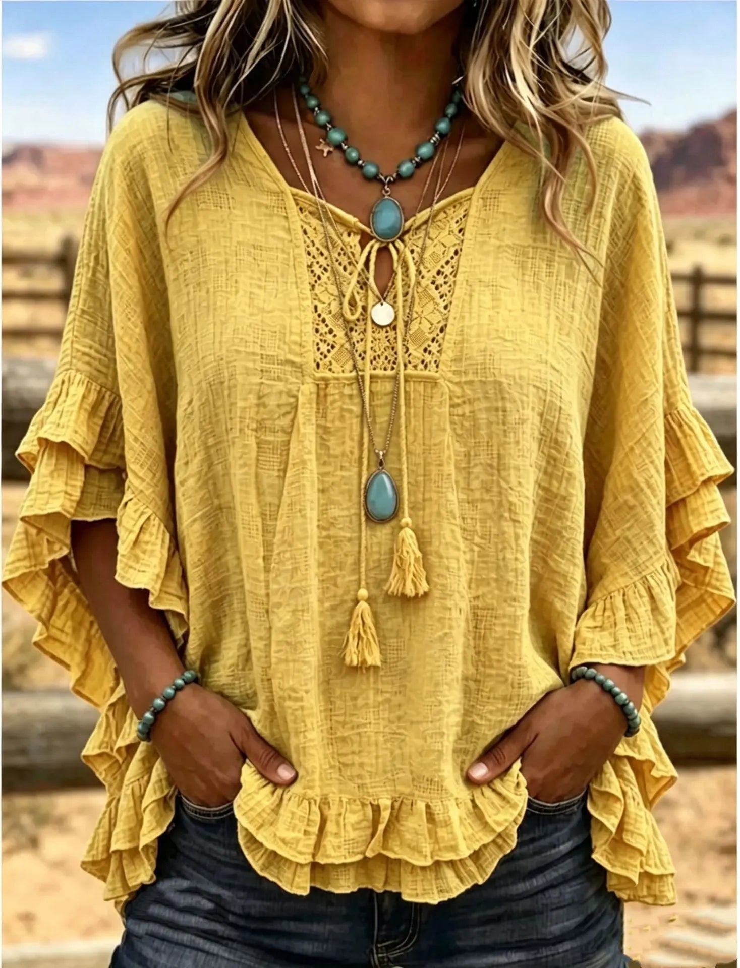 A woman wearing a loose, yellow bohemian-style blouse with tiered ruffle sleeves, lace-up neckline, and tassel detail, paired with dark jeans and layered turquoise necklaces, set against a blurred outdoor desert background.