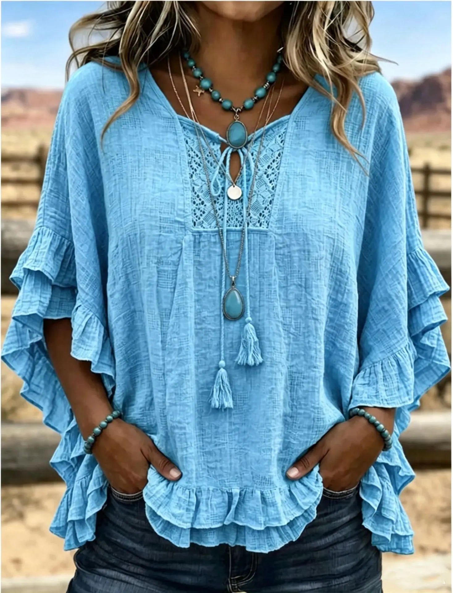 Blue ruffled blouse with lace details and tassels worn by a person, with a blurred natural background.