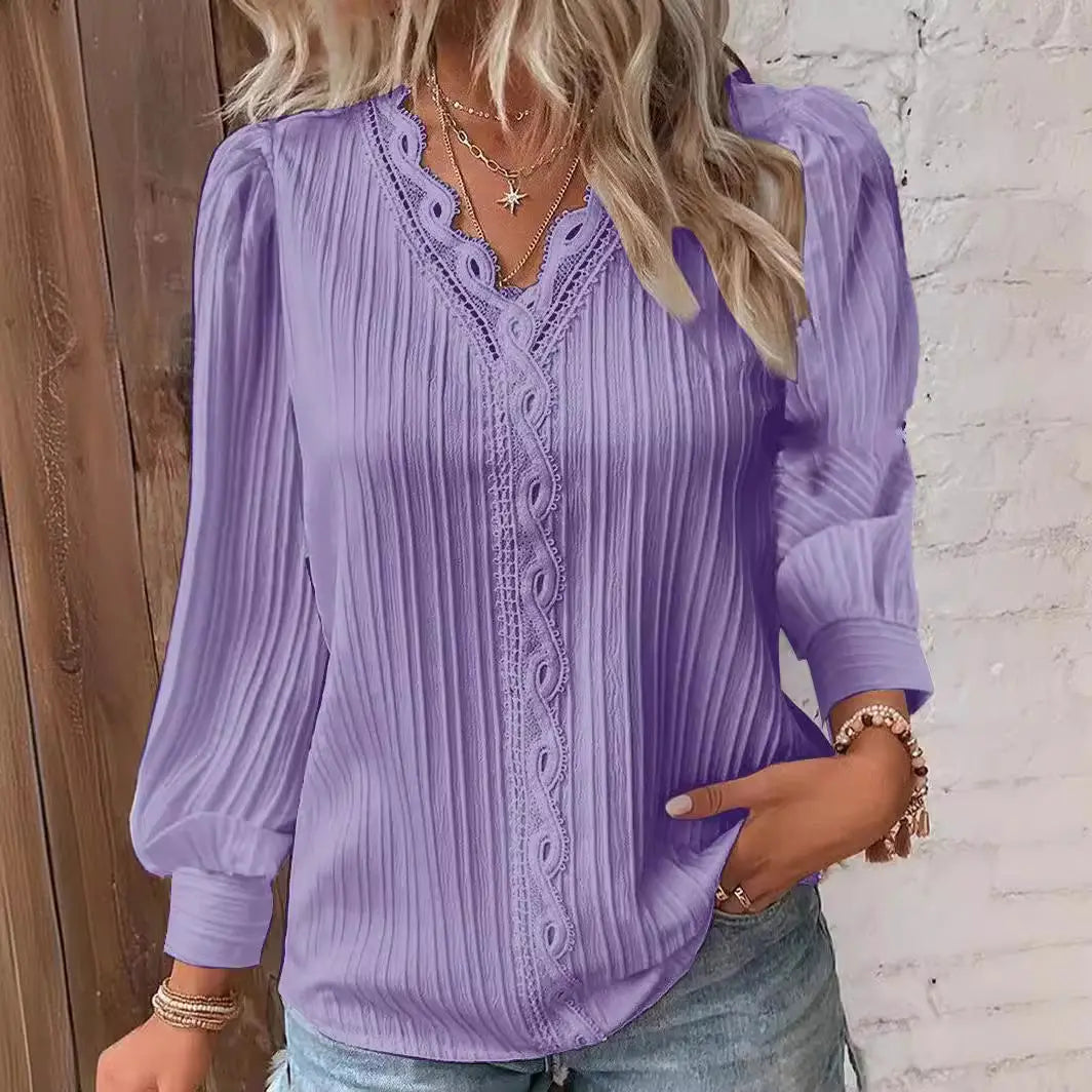 Woman wearing a purple blouse with lace details against a white brick wall.