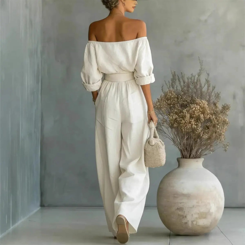Woman in a white off-shoulder top and pants holding a small bag, standing next to a large vase with dried plants.