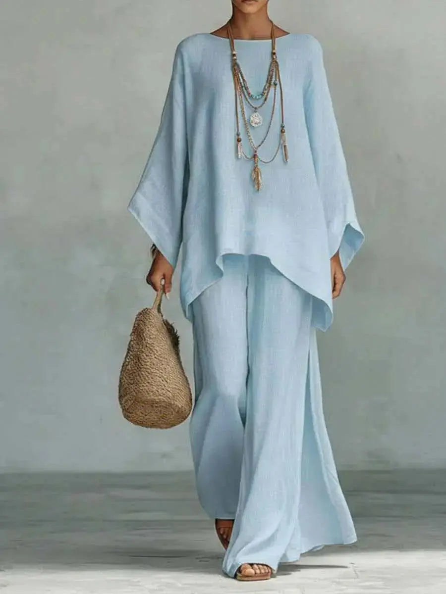 A model wearing a light blue linen two-piece set consisting of a wide-sleeve top and matching wide-leg pants, accessorized with layered necklaces and a woven straw bag against a gray studio background.