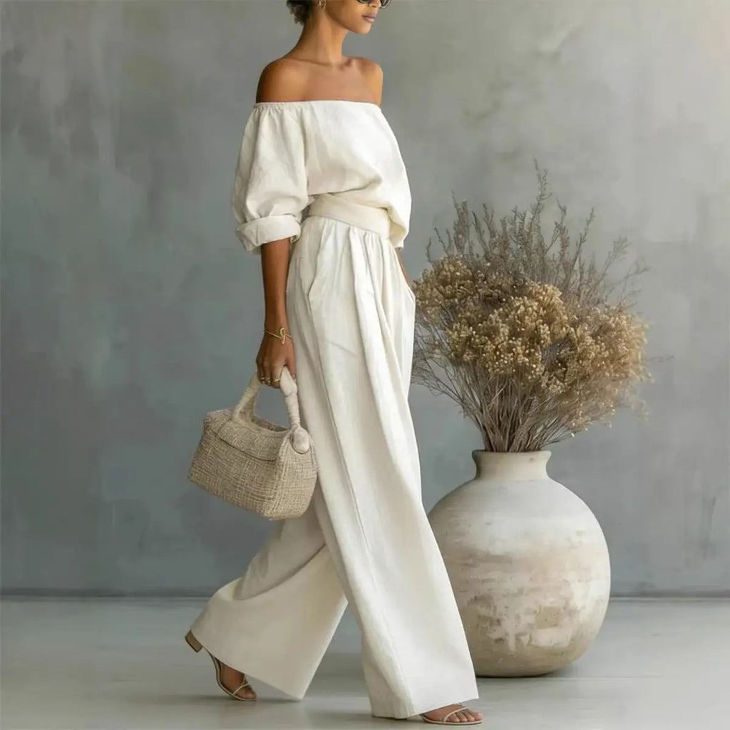 Woman in a white outfit holding a small bag, standing next to a large vase with dried flowers against a gray wall.