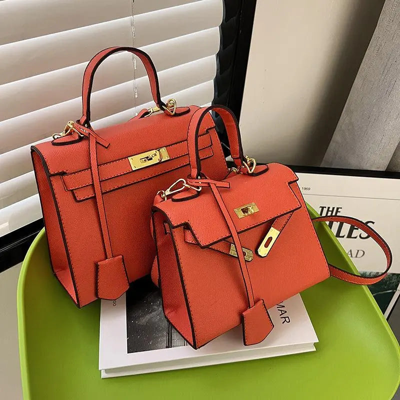 Two structured leather handbags in a warm coral-orange color with gold-tone hardware, displayed on a bright green tray indoors near window blinds.