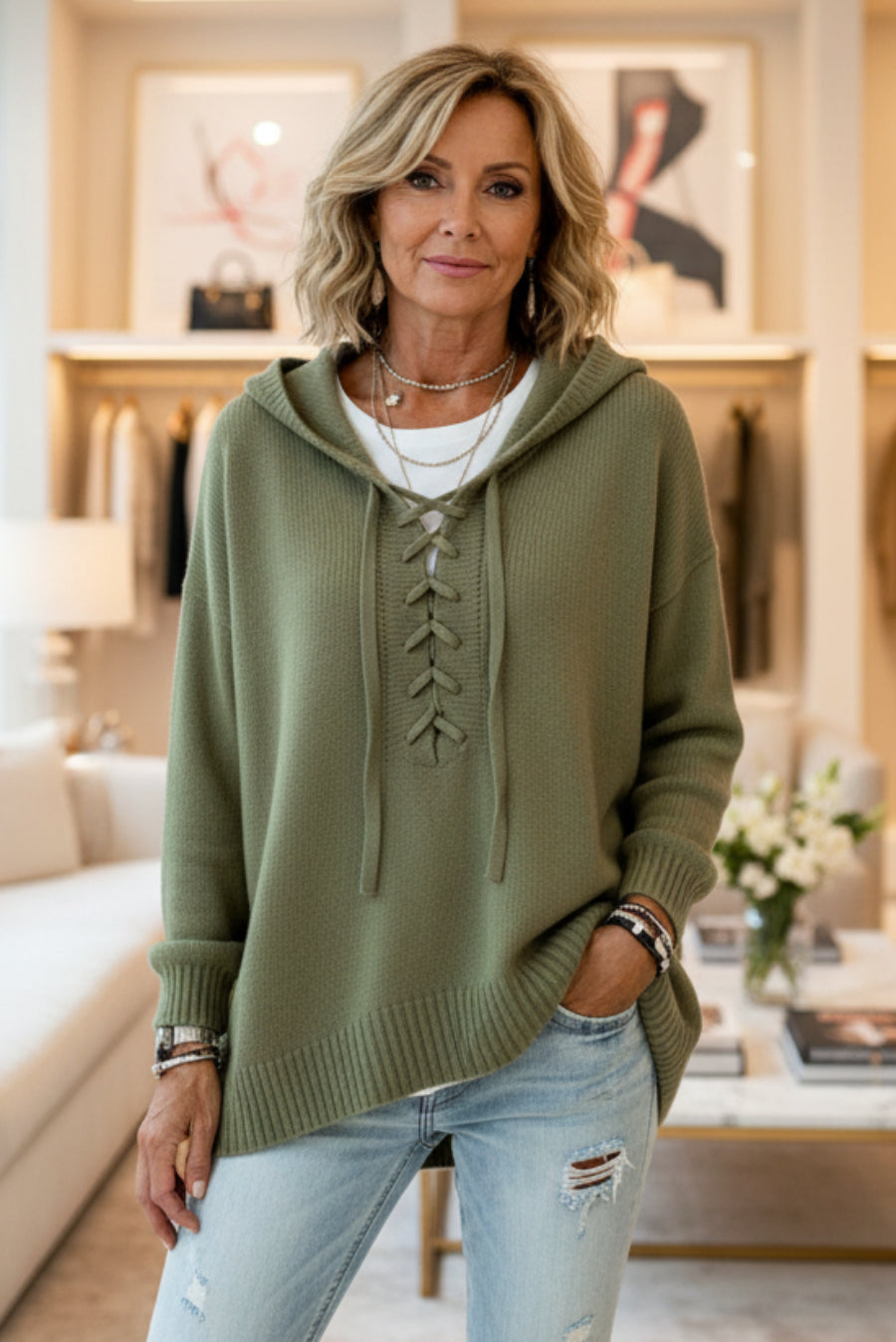 A woman wearing an olive green knit hooded sweater with lace-up front detailing, layered over a white top and paired with light blue jeans, standing in a softly lit boutique interior. Vicsxox.com