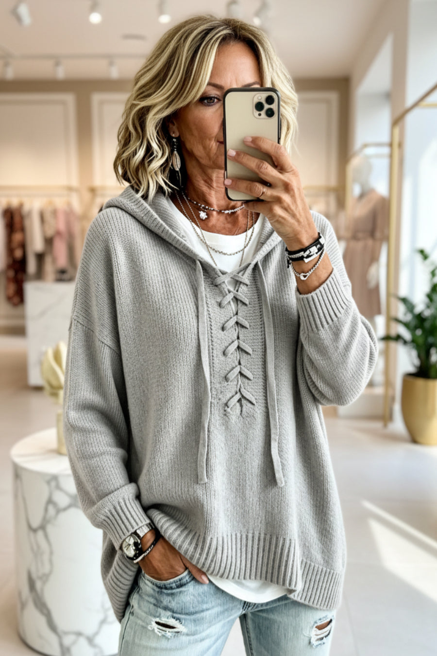 A woman takes a mirror selfie in a boutique while wearing a light gray ribbed knit hoodie with lace-up front detailing, a white t-shirt underneath, and distressed light-wash jeans. Vicsxox.com
