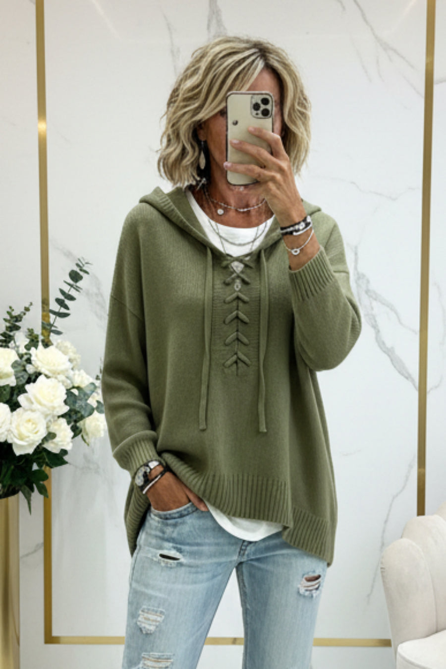 A woman takes a mirror selfie wearing an olive green knit hooded sweater with lace-up front detailing, paired with light blue distressed jeans and a white undershirt. The background is a clean interior with white marble walls, gold trim, and a vase of white roses. Vicsxox.com