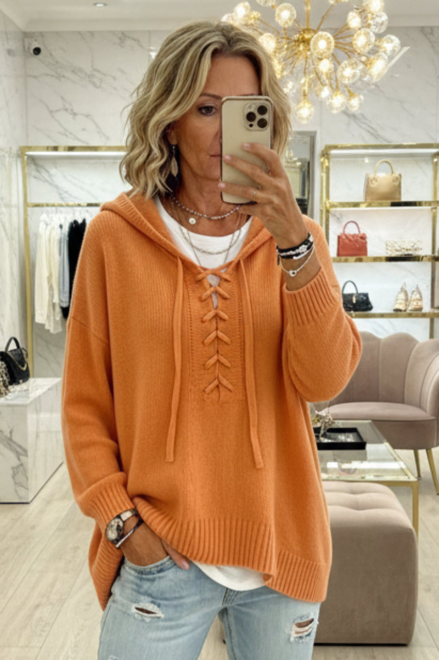 A woman takes a mirror selfie in a boutique wearing an oversized orange knit hoodie with lace-up front detailing, light blue distressed jeans, and layered necklaces. Vicsxox.com