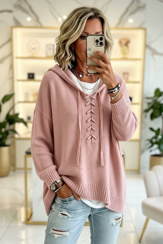 A woman takes a mirror selfie wearing a dusty pink lace-up hooded sweater over a white t-shirt and light-wash distressed jeans, set in a bright, modern room with marble floors and shelving. Vicsxox.com