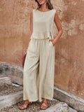A woman models a sleeveless, light beige linen jumpsuit with a ruffled waist and wide-leg pants, paired with brown leather sandals, standing against a textured stucco wall. Vicsxox.com