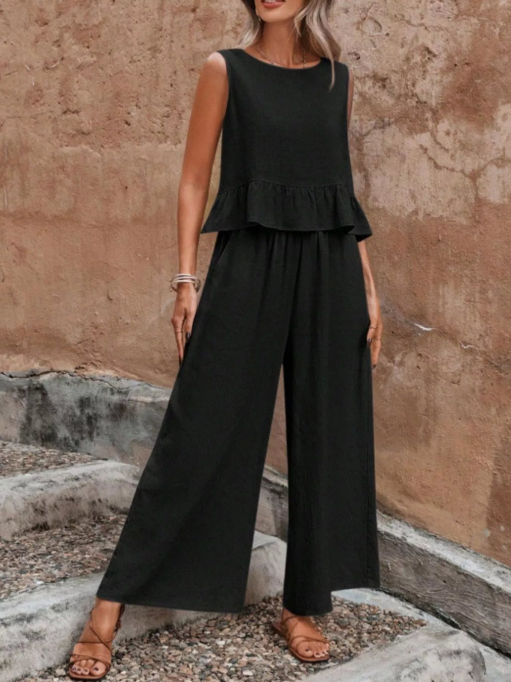 Woman wearing a black peplum jumpsuit standing against a textured wall. Vicsxox.com