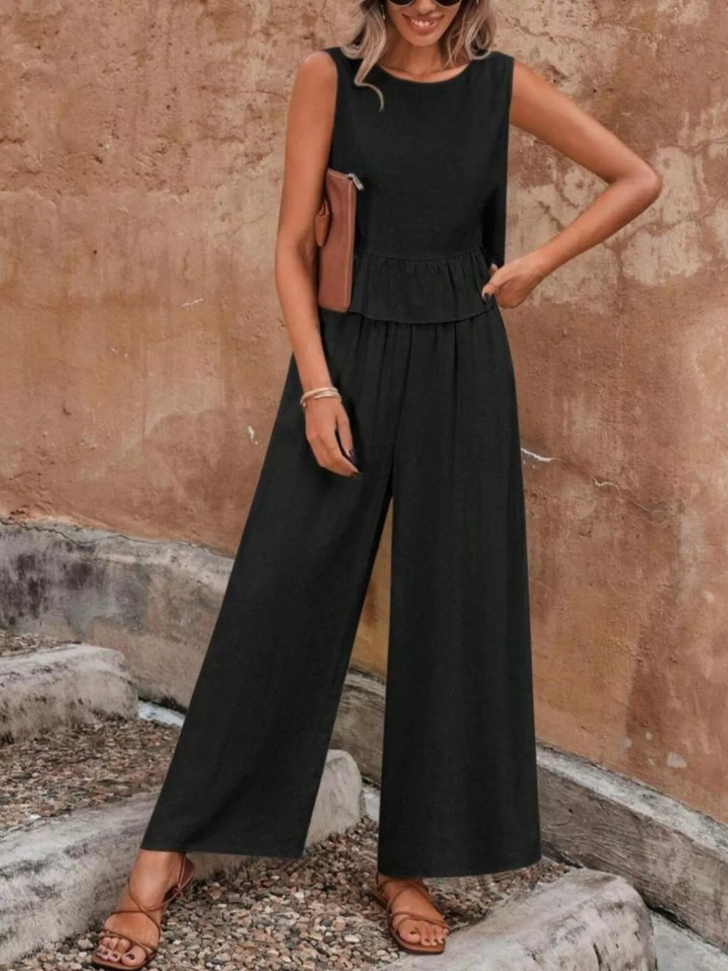Woman wearing a black sleeveless jumpsuit against a textured wall. Vicsxox.com