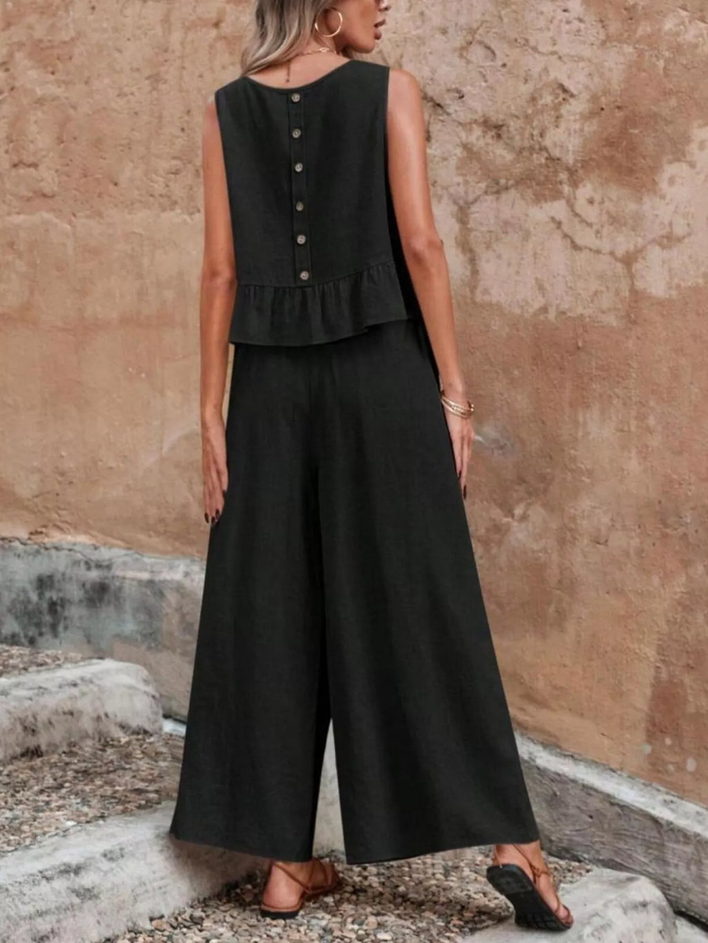 Woman wearing a black sleeveless top and wide-leg pants against a textured wall. Vicsxox.com