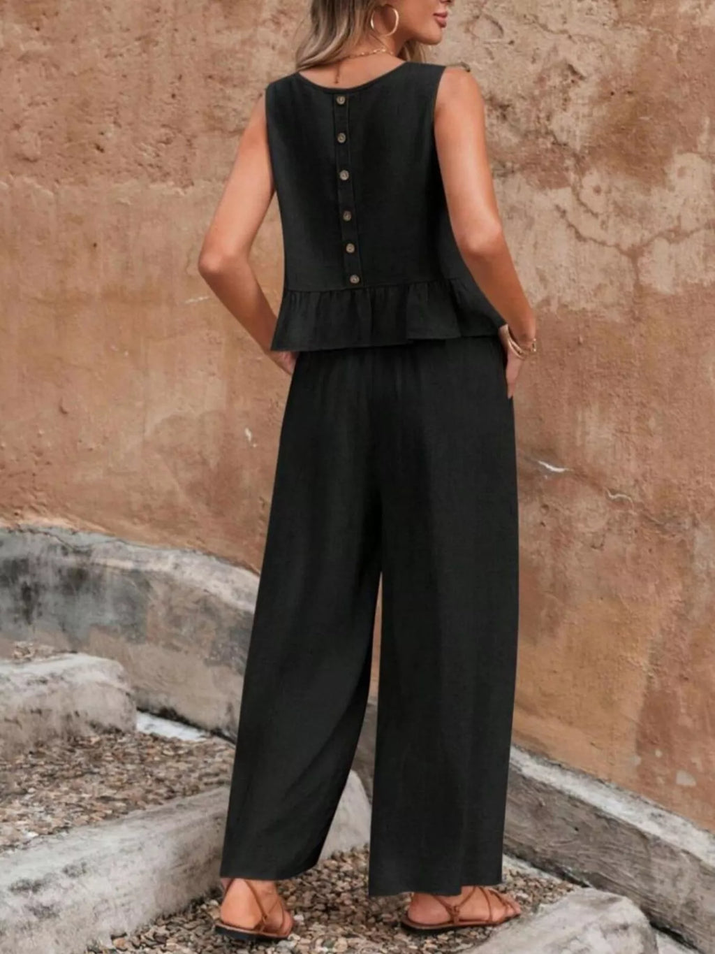 Person wearing a black sleeveless top and wide-leg pants against a textured wall. Vicsxox.com