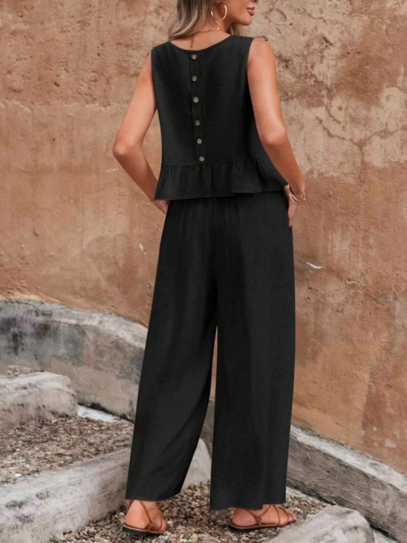 Person wearing a black sleeveless top and wide-leg pants against a textured wall. Vicsxox.com