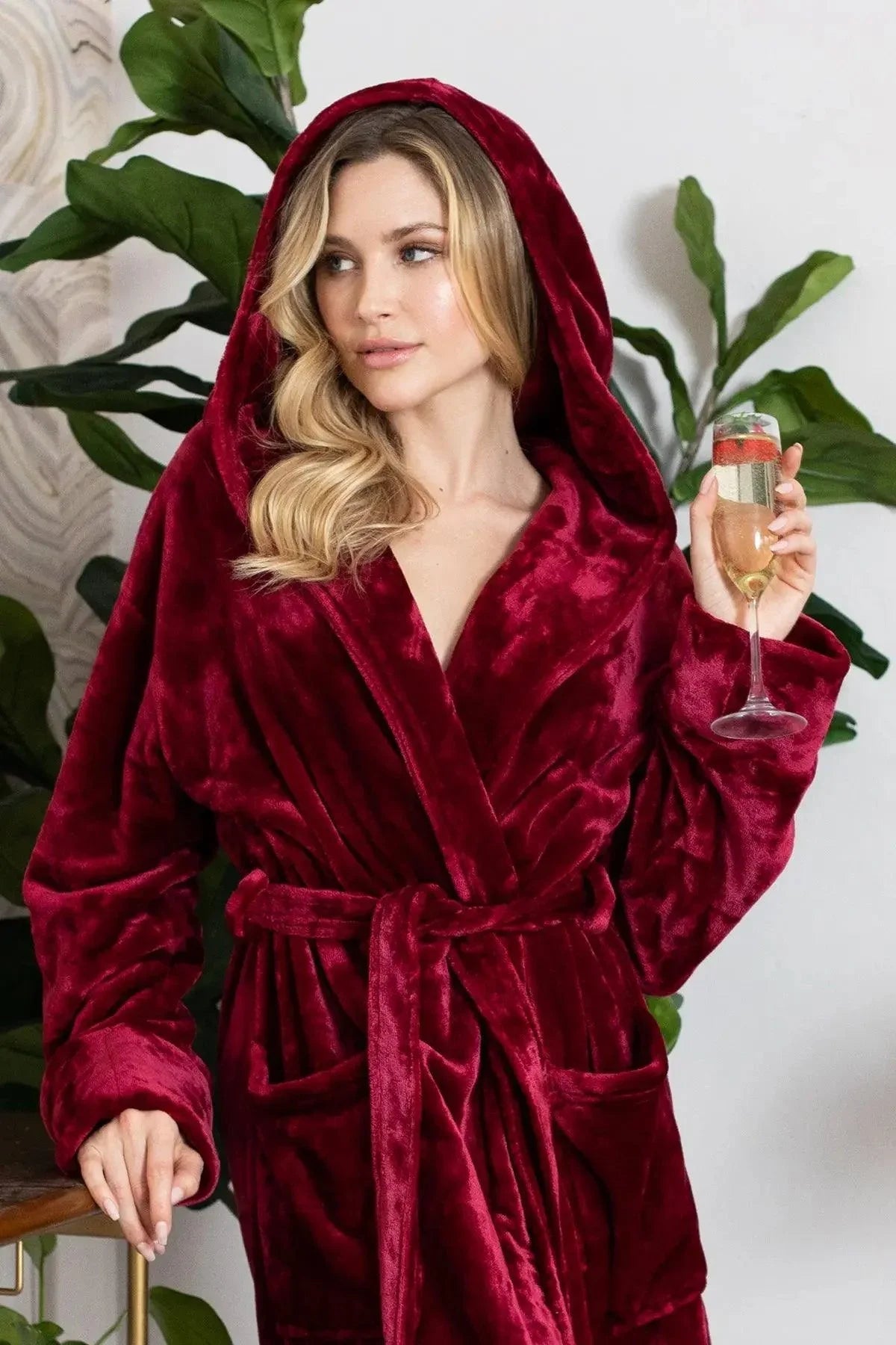 Woman wearing a red velvet robe holding a glass of wine against a white background with green plants. Vicsxox.com