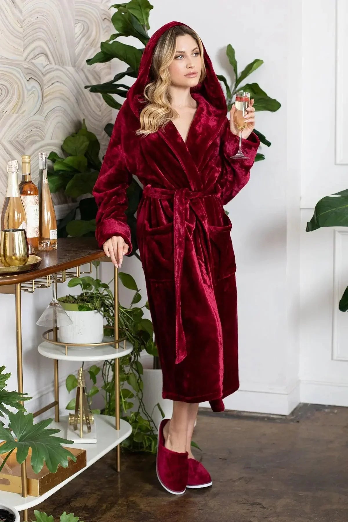Woman in a red robe holding a glass in a stylish room with plants and decor. Vicsxox.com