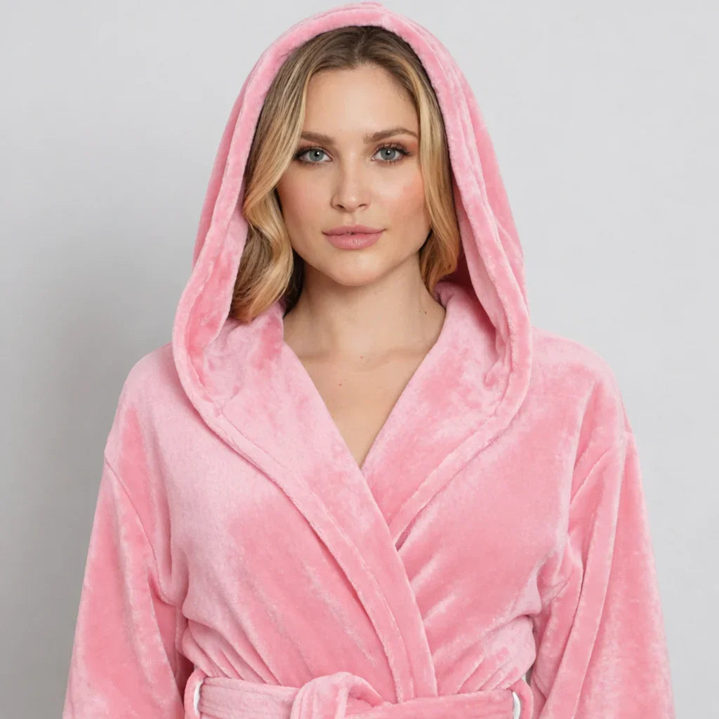 A woman wearing a plush pink hooded robe with a tie belt, shown from the chest up against a light gray studio background. Vicsxox.com