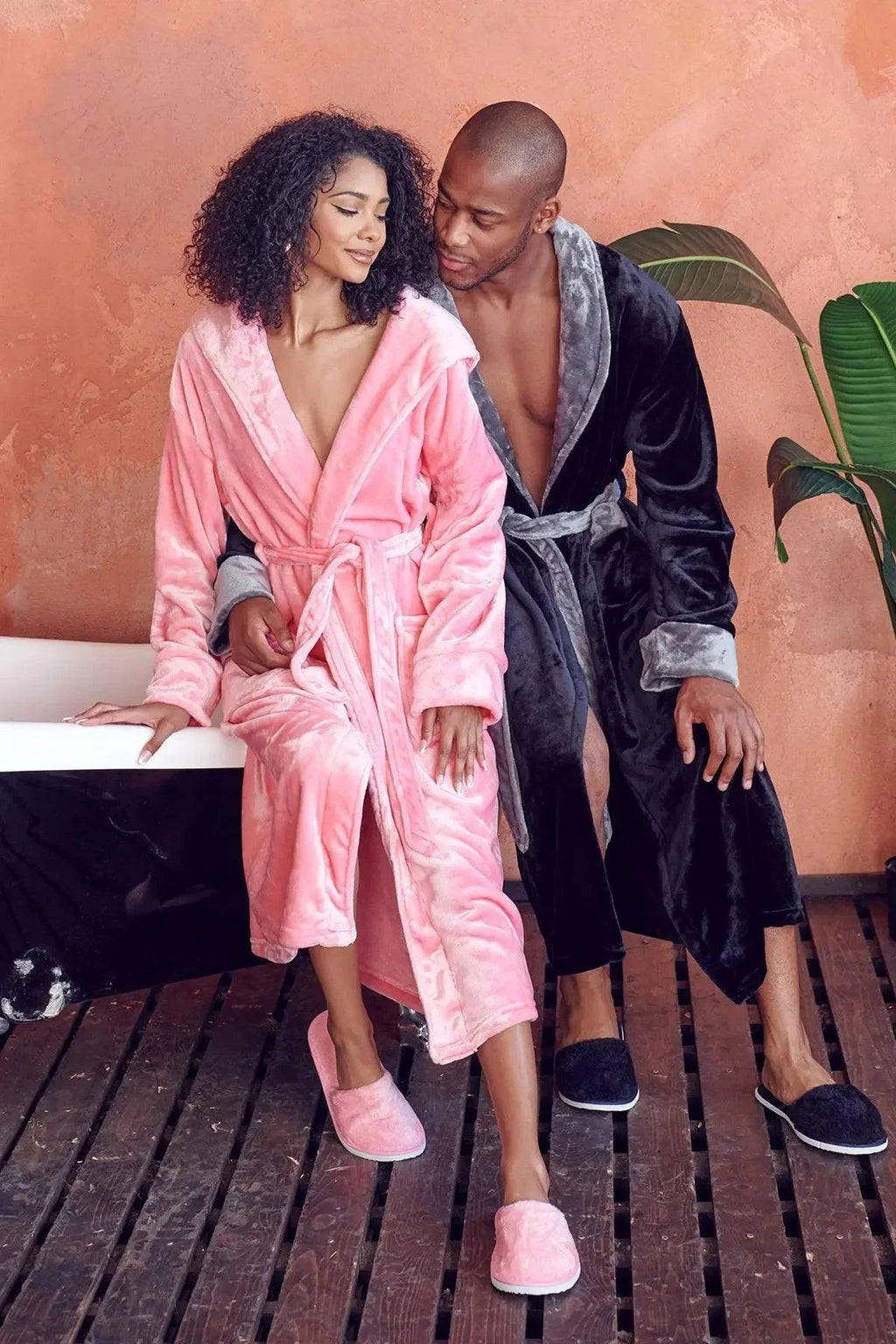 Two people in plush robes sitting on a wooden floor with a pink wall in the background. Vicsxox.com