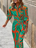 A woman wearing a green and orange abstract patterned wrap dress with three-quarter sleeves, walking outdoors on a stone sidewalk next to a stone wall.
