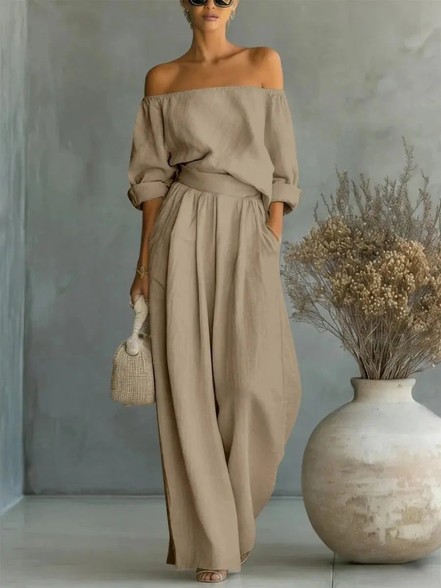 Woman wearing a beige off-shoulder top and wide-leg pants, standing next to a vase with dried flowers.