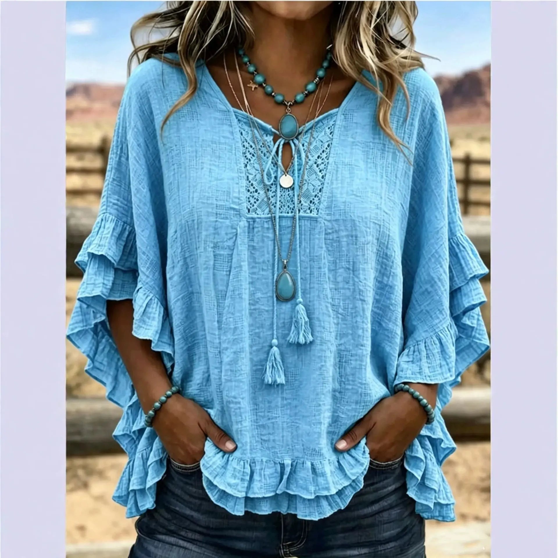 A woman wearing a light blue bohemian-style blouse with layered ruffle sleeves, a lace V-neckline, and a tassel tie detail, styled with turquoise jewelry against a rustic outdoor background.