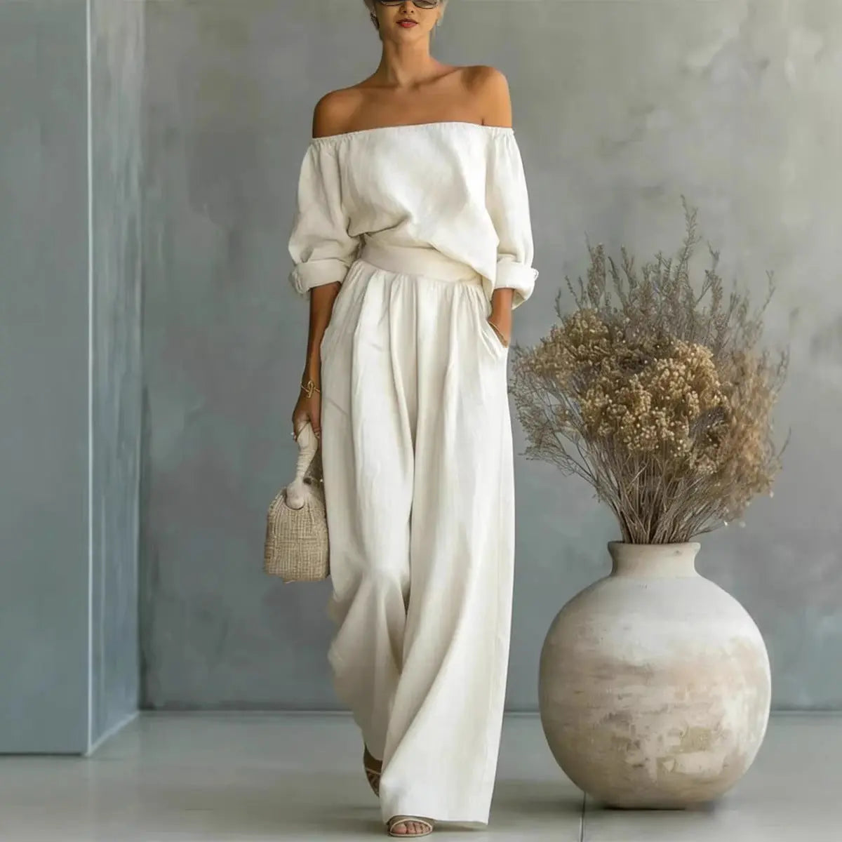 Woman in a white off-shoulder top and wide-leg pants standing next to a vase with dried flowers.