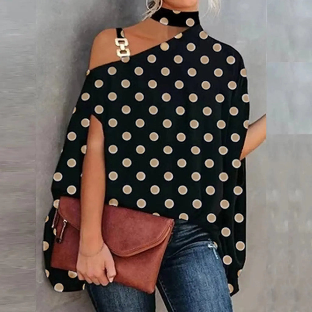 Woman wearing a black polka dot top with cut-out shoulders, holding a brown clutch against a gray background.