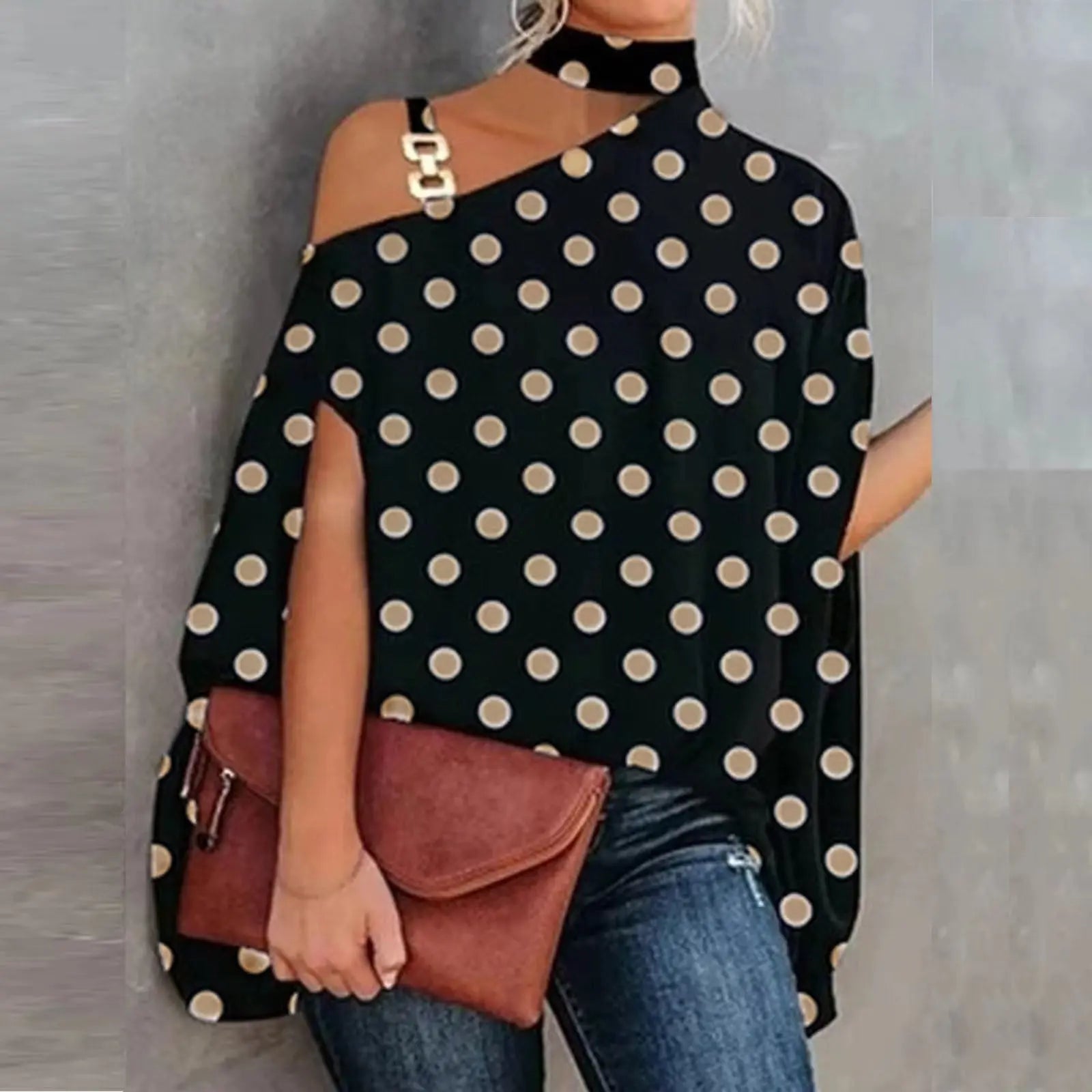 Woman wearing a black polka dot top with cut-out shoulders, holding a brown clutch against a gray background.