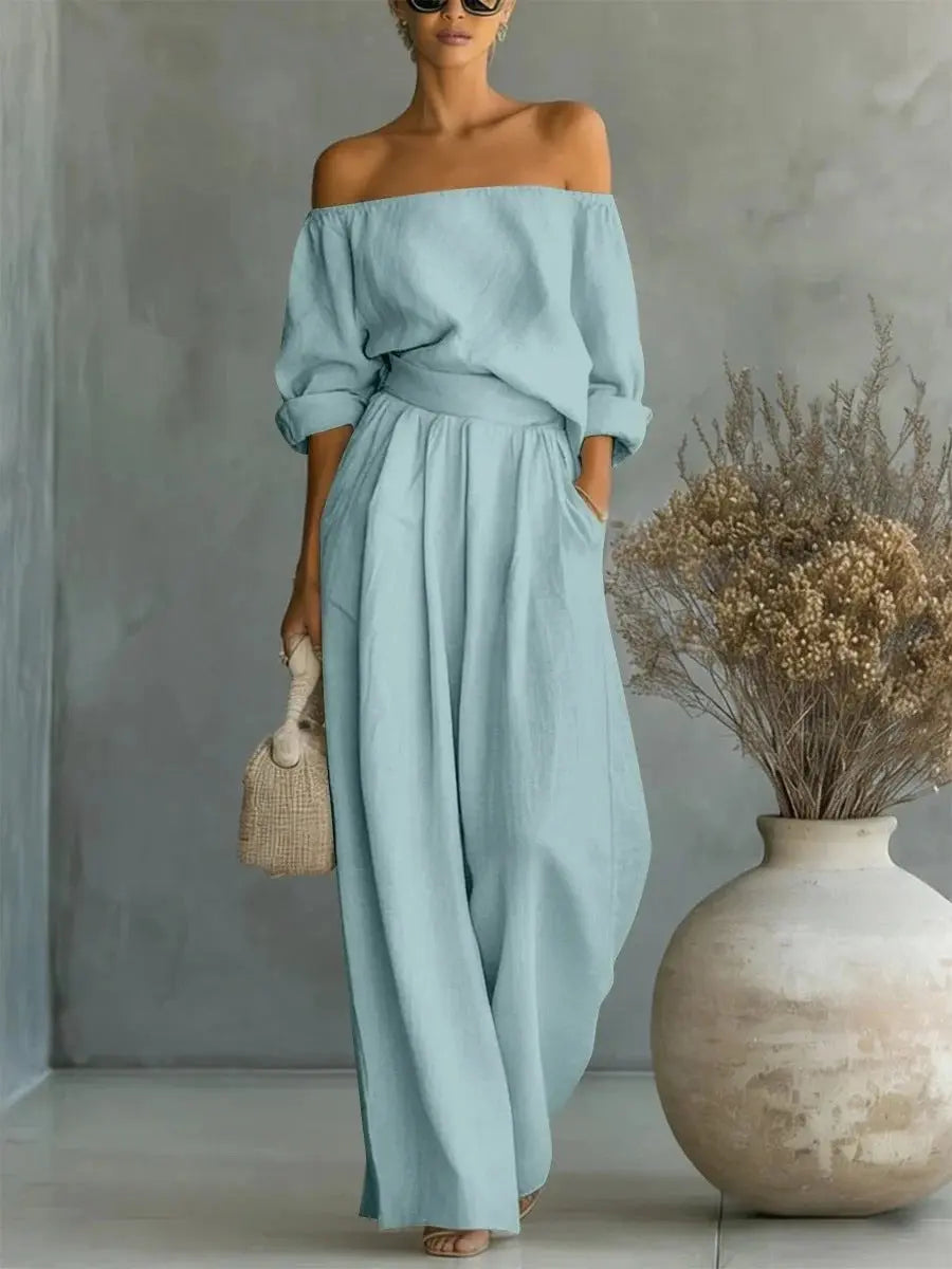Woman wearing a light blue off-shoulder dress standing against a neutral background with a vase and dried plants.