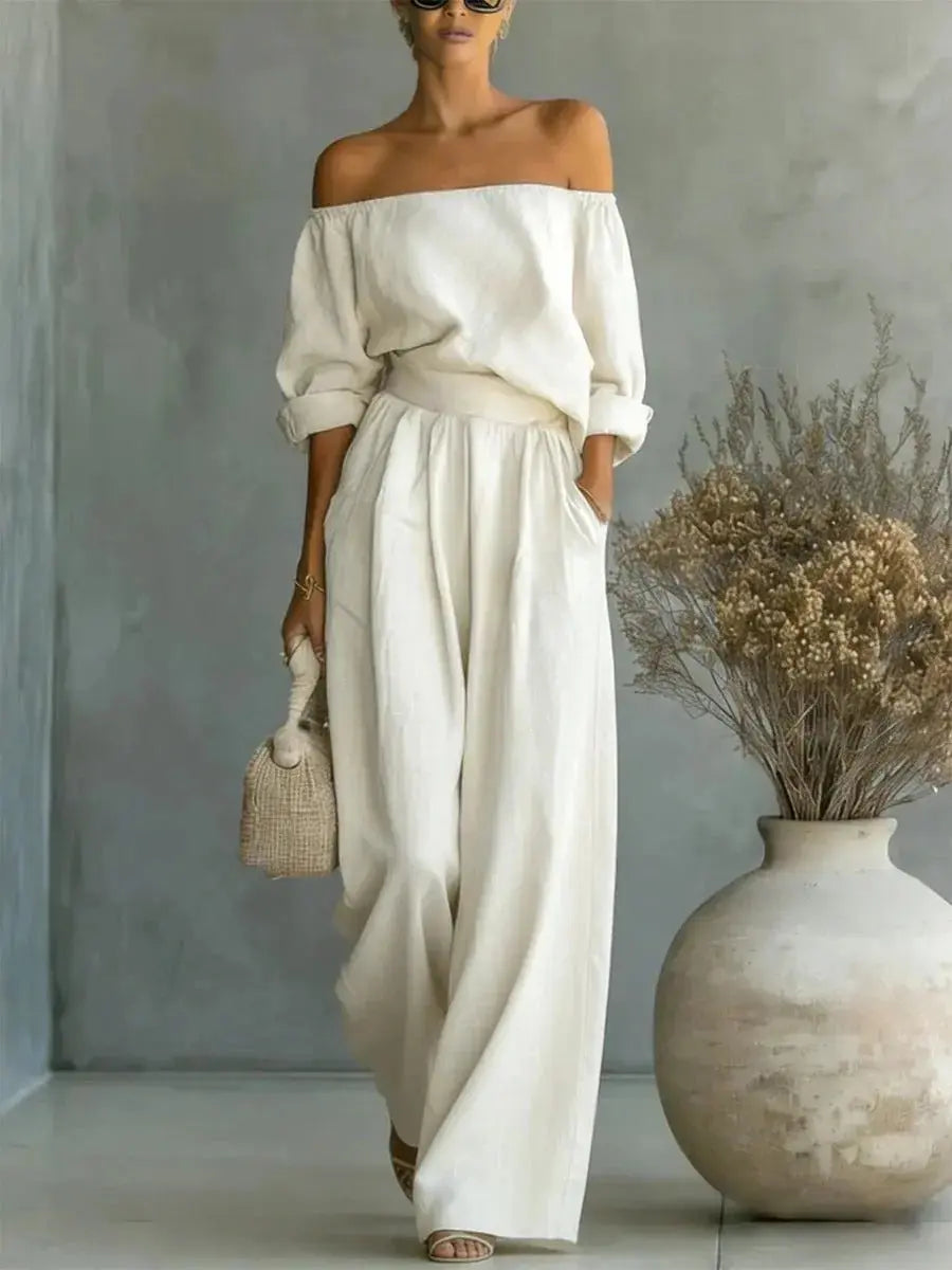 Woman in a white off-shoulder outfit standing next to a vase with dried flowers against a gray wall.