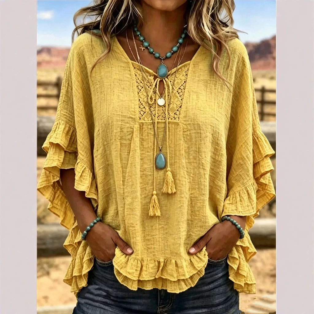 Yellow blouse with ruffled sleeves and a necklace with a blue pendant, set against a blurred outdoor background.