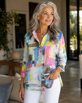 A woman with long gray hair wearing a colorful abstract print button-down shirt and white pants, standing in a modern indoor setting. Vicsxox.com